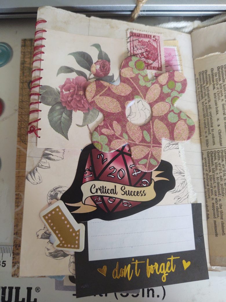 A junk journal page made up of red flower themed ephemera, a D20 critical success sticker and a don't forget post-it note